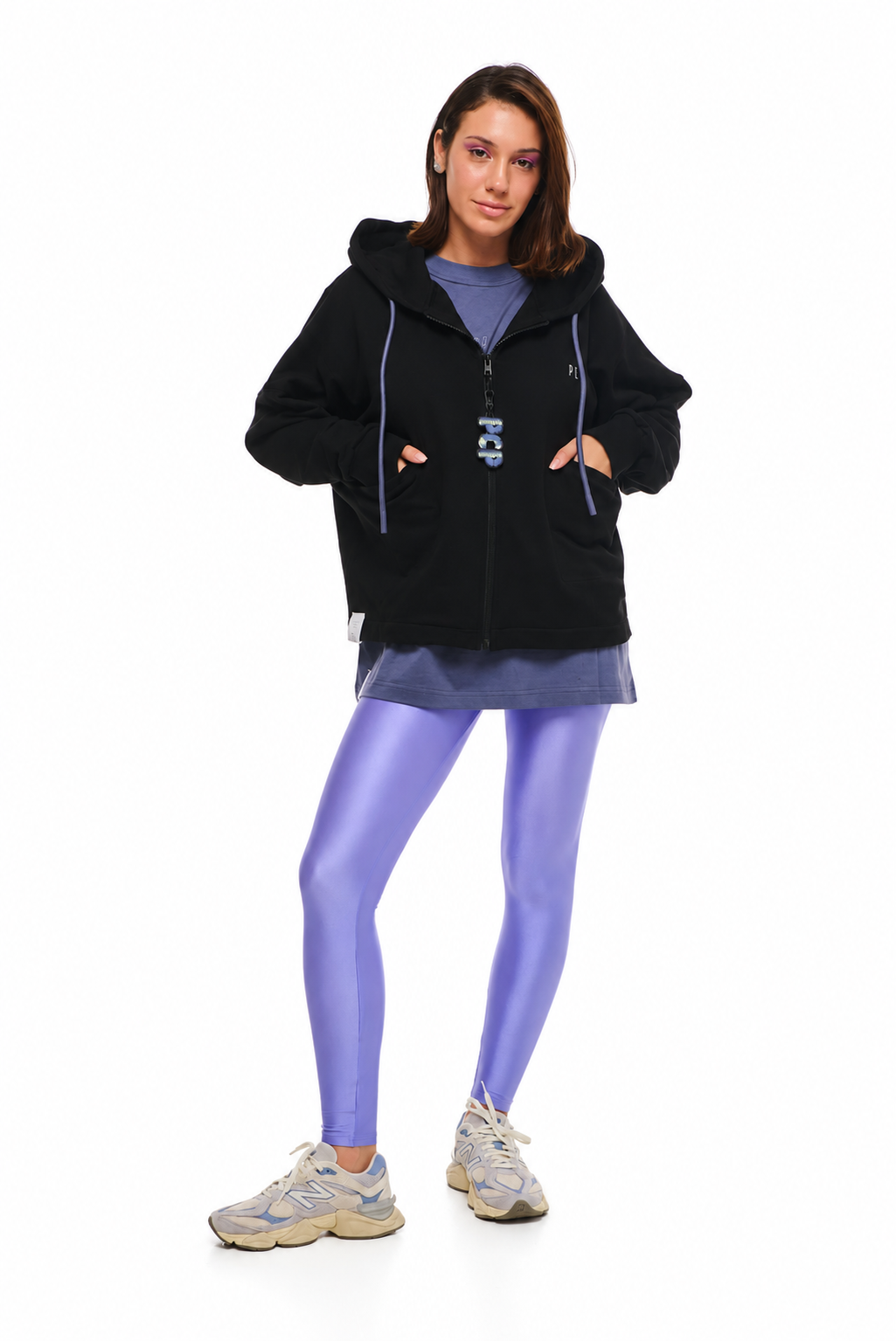A woman modeling a black zip-up hoodie with purple drawstrings over a purple top and skirt, paired with shiny purple leggings and gray sneakers, against a plain white background.