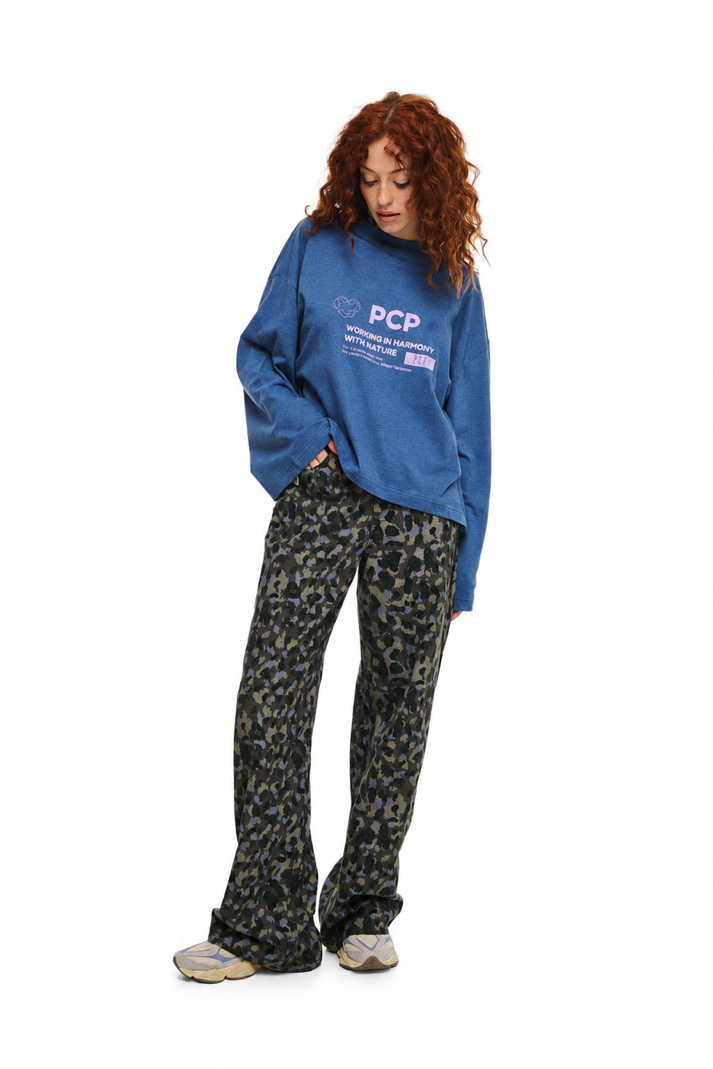 A model with curly red hair wearing a blue sweatshirt with 'PCP' printed on the chest and dark camouflage pants, standing against a plain white background.