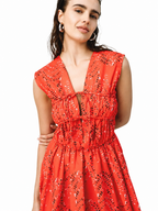 A woman wearing a sleeveless red dress with a black abstract pattern, featuring a deep V-neckline, tie-front detail, and a gathered smocked waist, set against a plain white background.