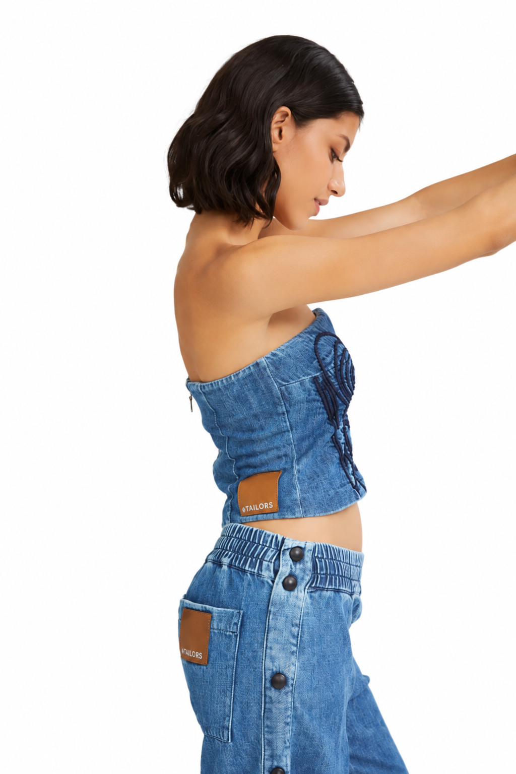 A woman in side profile wearing a blue denim strapless crop top and matching high-waisted jeans with brown leather brand patches, set against a plain white background.