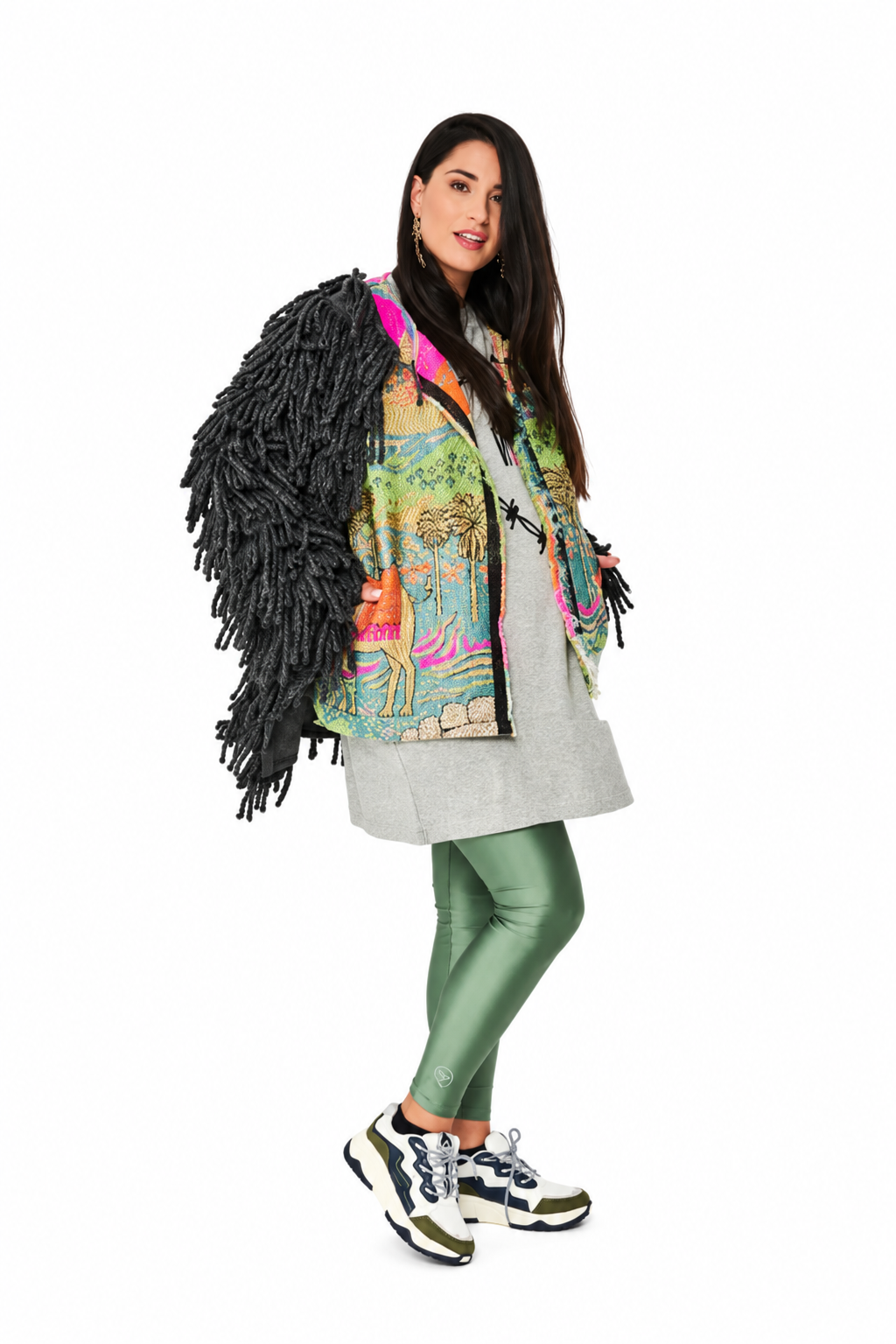 A woman modeling a unique outerwear piece featuring a colorful, illustrated print on the front, a light gray tunic underneath, and a large black shaggy fringe shoulder flap, paired with metallic green leggings and sneakers against a white background.