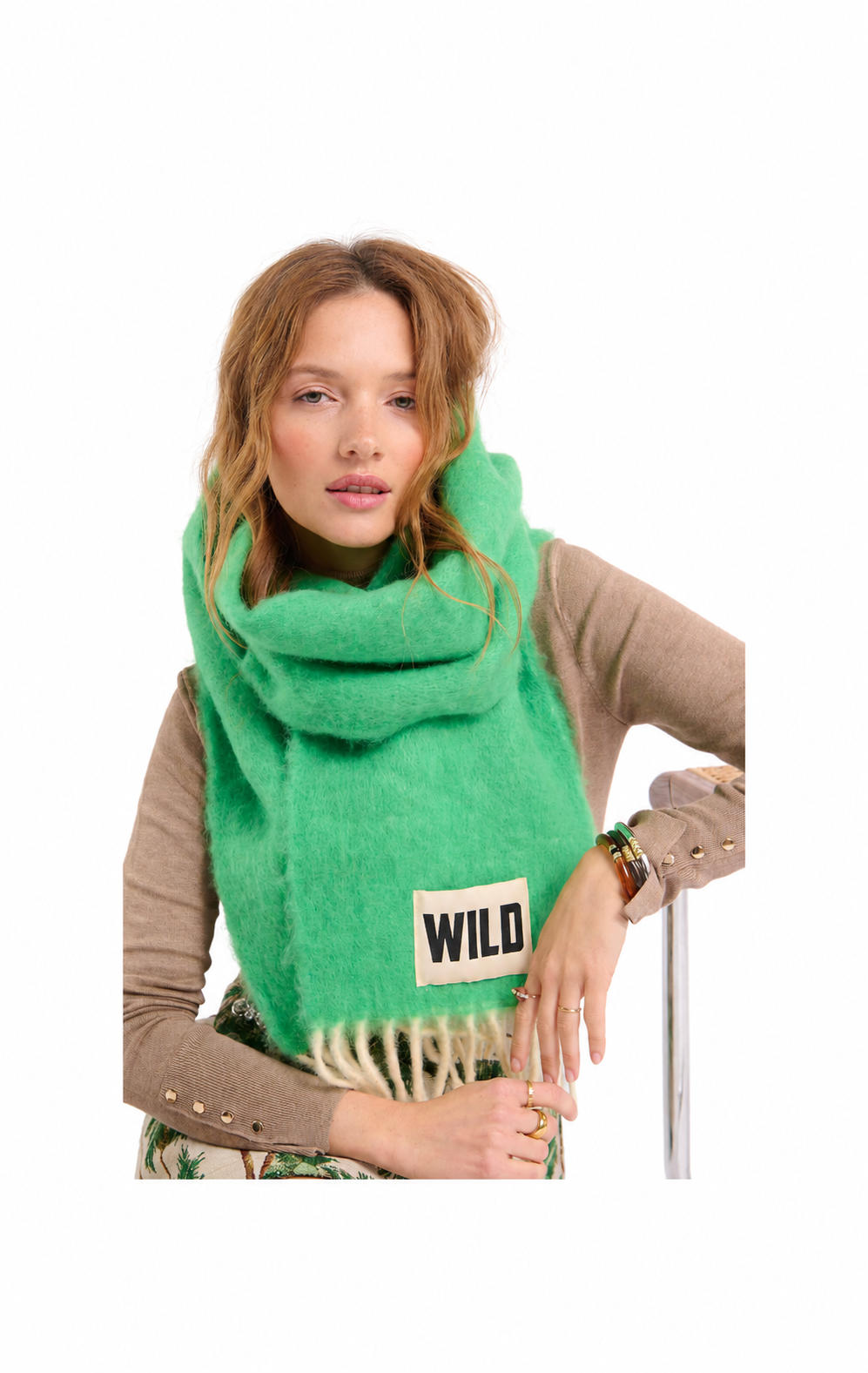 A woman wearing a bright green fuzzy knit scarf with cream fringe and a small beige label that reads 'WILD', paired with a brown top against a plain white background.