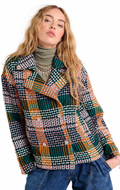 A woman wearing a multicolor plaid tweed blazer with green, orange, white, and black patterns, styled over a green turtleneck and blue jeans against a white background.