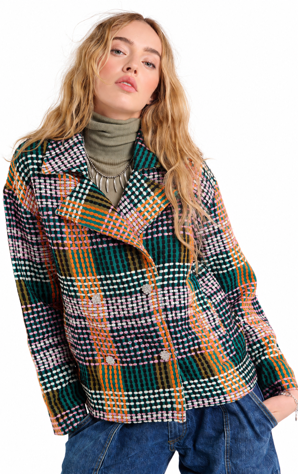 A woman wearing a multicolor plaid tweed blazer with green, orange, white, and black patterns, styled over a green turtleneck and blue jeans against a white background.