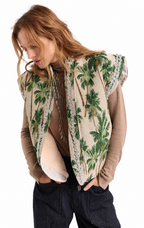 A woman wearing a sleeveless beige vest with a green palm tree pattern and silver trim, layered over a brown long-sleeve top and dark jeans, against a plain white background.