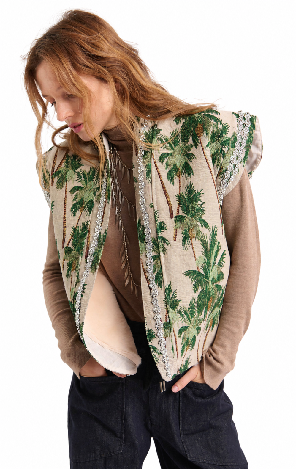 A woman wearing a sleeveless beige vest with a green palm tree pattern and silver trim, layered over a brown long-sleeve top and dark jeans, against a plain white background.