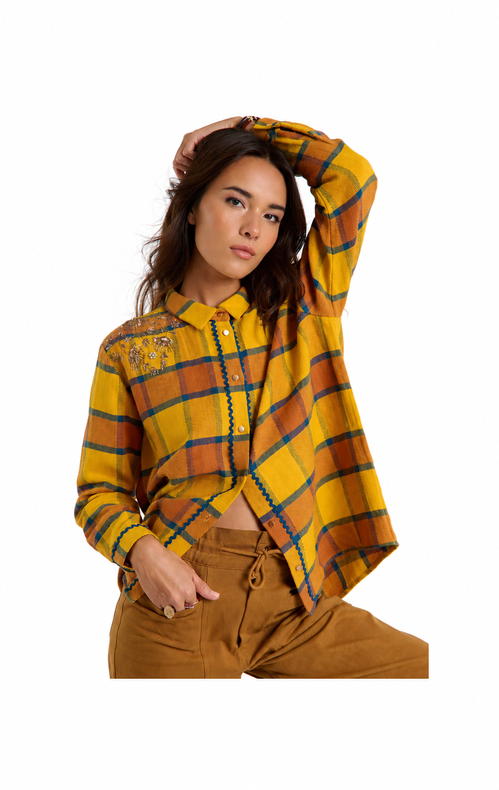 A woman modeling a cropped, long-sleeve plaid button-up shirt in mustard yellow, orange, and dark green, paired with brown pants, against a solid white background.
