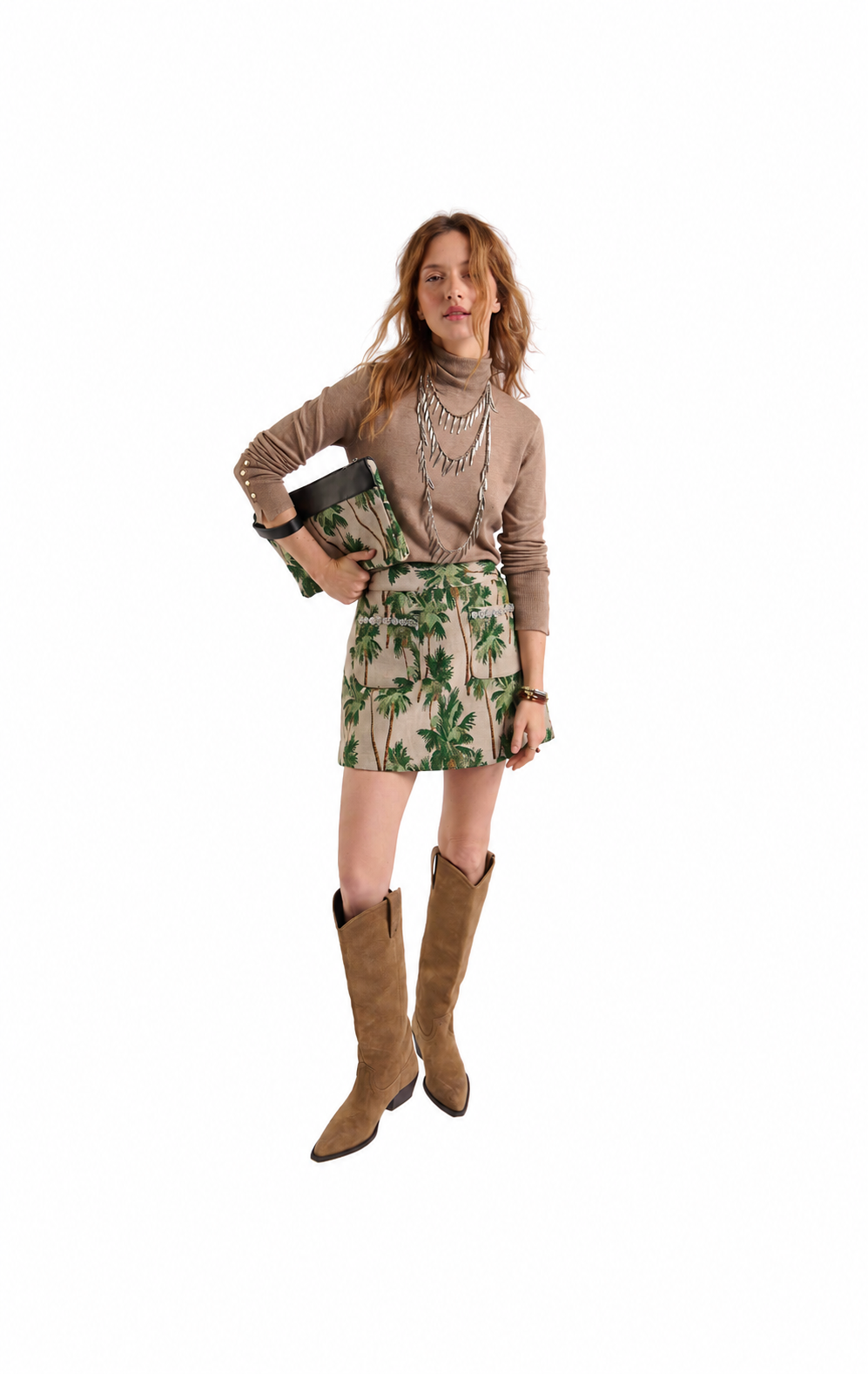 A model stands against a white background wearing a beige long-sleeve top, a beige mini skirt with a green tropical leaf print, brown suede cowboy boots, and holding a dark green clutch.