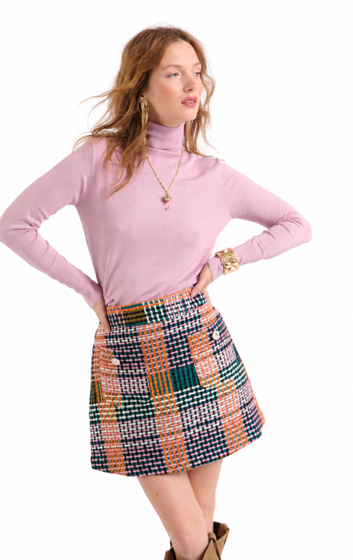 A woman modeling a light pink long-sleeve turtleneck sweater and a colorful patchwork tweed mini skirt with orange, green, white, and navy plaid patterns, set against a plain white background.