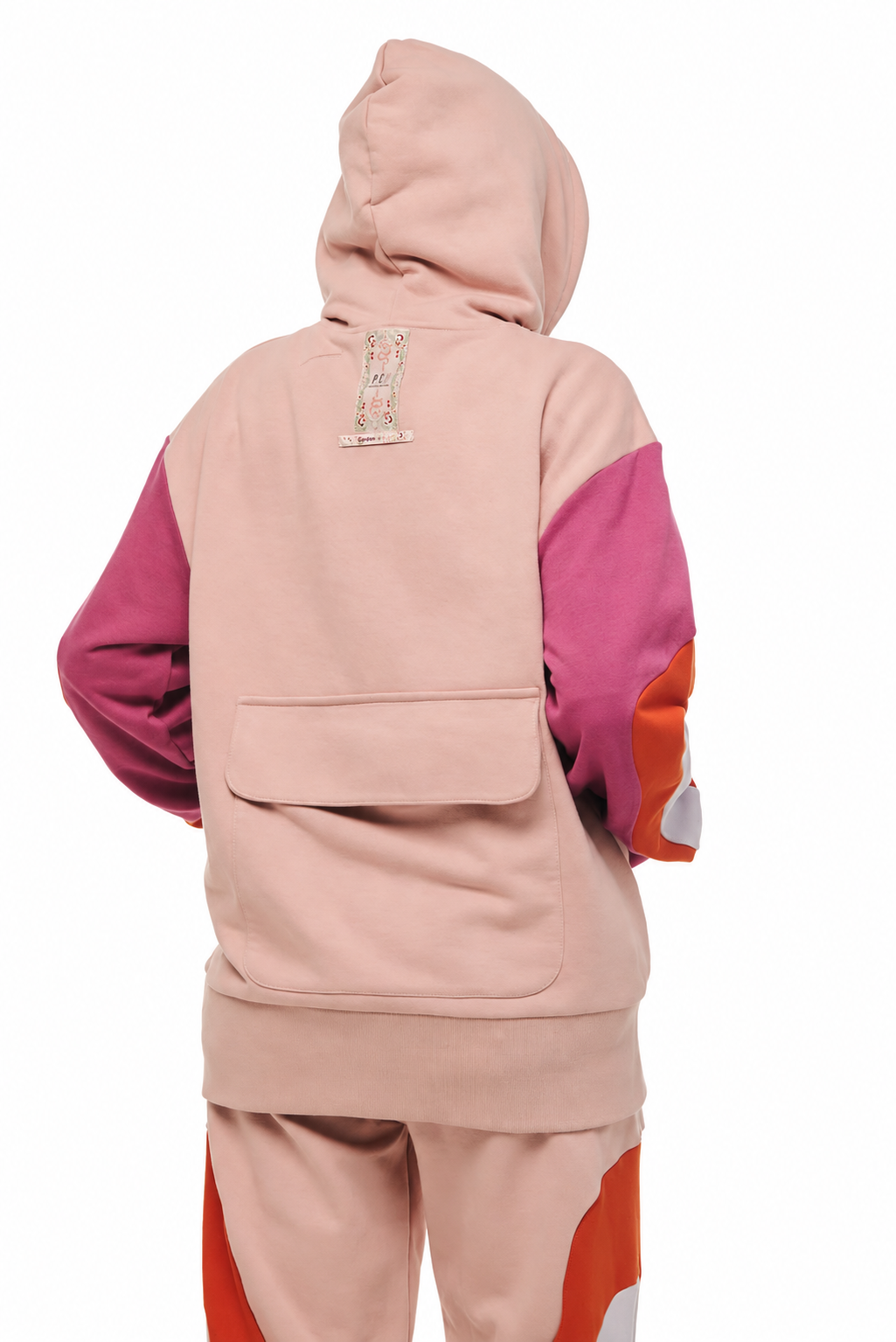 A person shown from the back wearing a color-blocked hoodie and matching sweatpants. The hoodie is primarily dusty pink with magenta sleeves and orange side panels, with a small embroidered logo on the back neck.