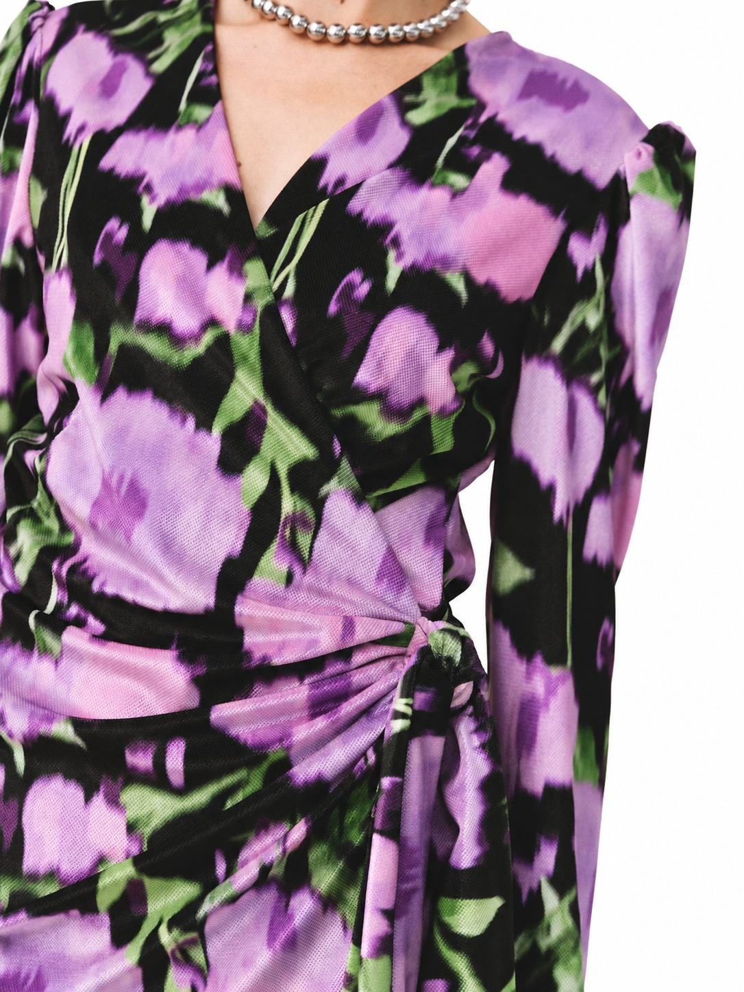 A close-up of a long-sleeve wrap dress featuring a bold purple, pink, and green floral pattern over a black base, styled with a silver beaded necklace.