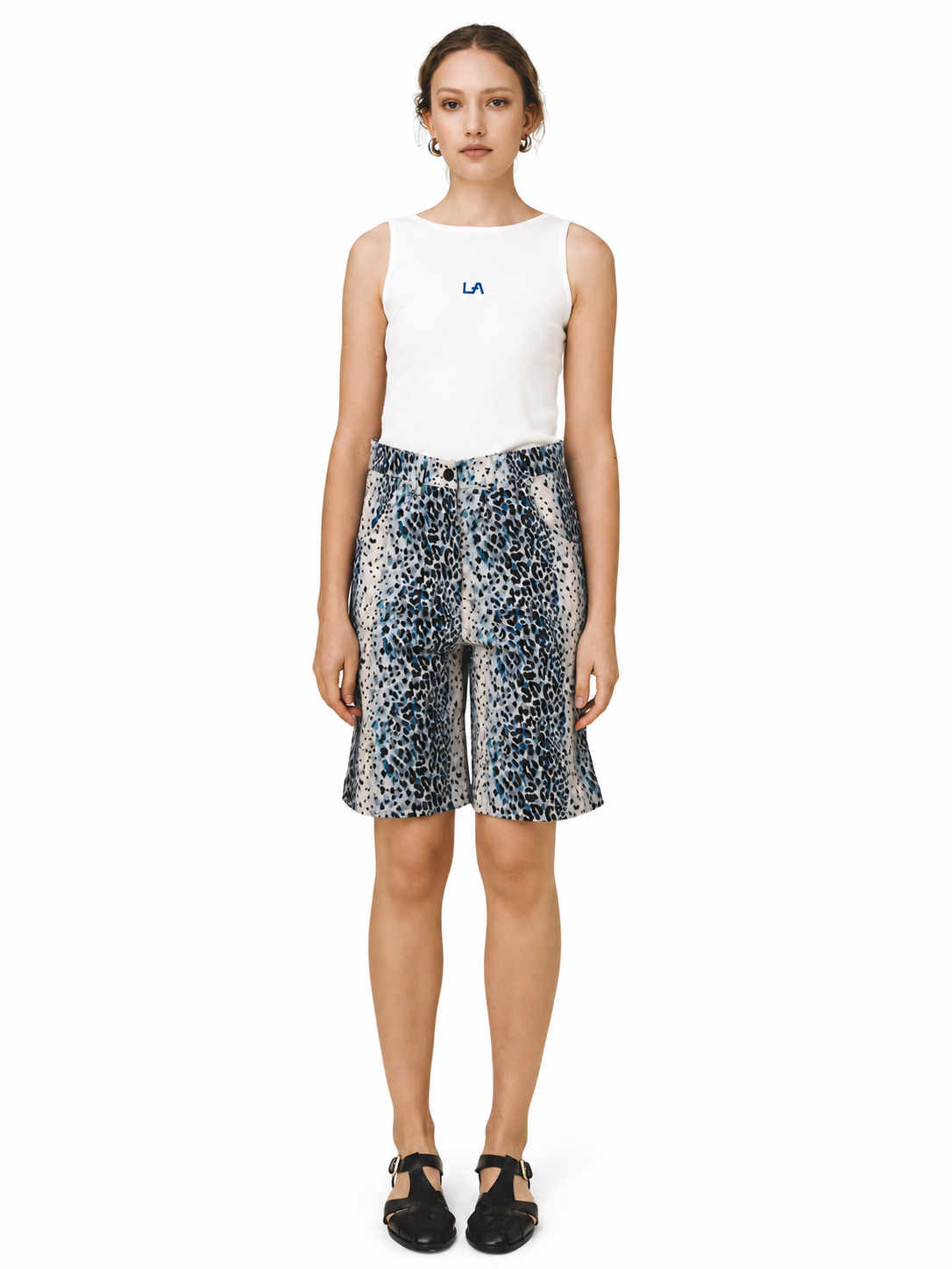 A model stands against a white studio background wearing a white sleeveless top with a small blue LA logo on the chest, paired with blue, black, and white leopard-print shorts, and black Mary Jane-style shoes.