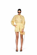 A model stands against a white studio background wearing a light yellow patterned button-up shirt and matching shorts, black sunglasses, black strappy heels, and holding a matching yellow patterned clutch with a gold chain strap.