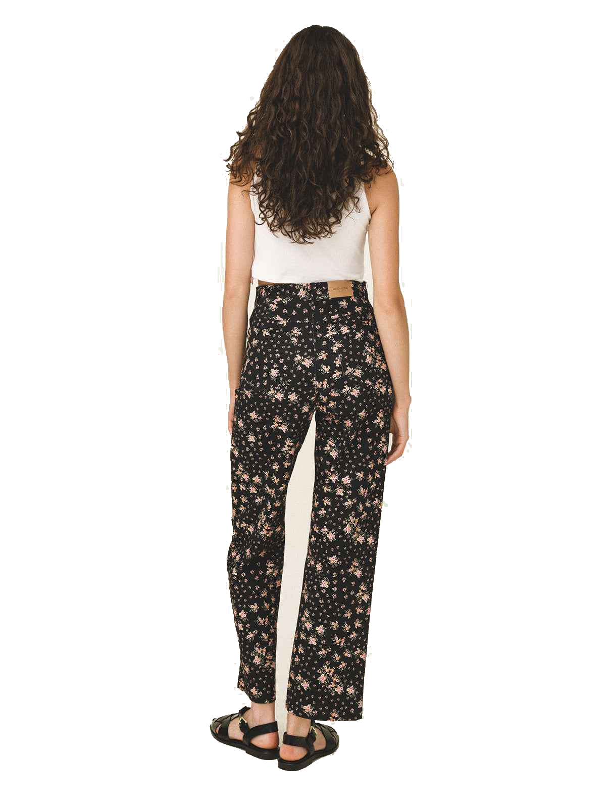 A woman viewed from the back wearing black wide-leg pants with a beige floral pattern and a white side stripe, paired with a white sleeveless top and black sandals, on a white background.