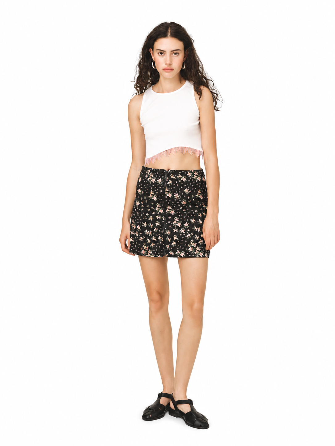 A model stands against a solid white background wearing a white sleeveless crop top, a black mini skirt with a beige floral pattern, and black Mary Jane shoes.