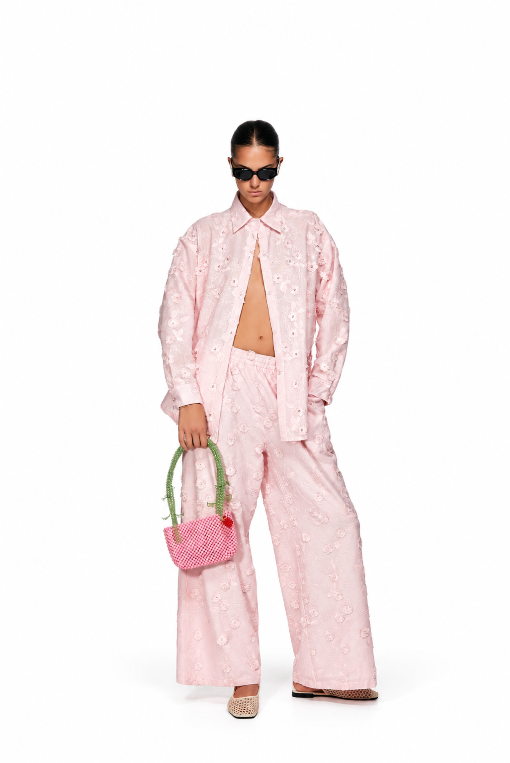 A model wearing a light pink embroidered two-piece set consisting of a long-sleeve button-up shirt and matching wide-leg trousers, holding a small pink patterned handbag with green handles against a solid white background.