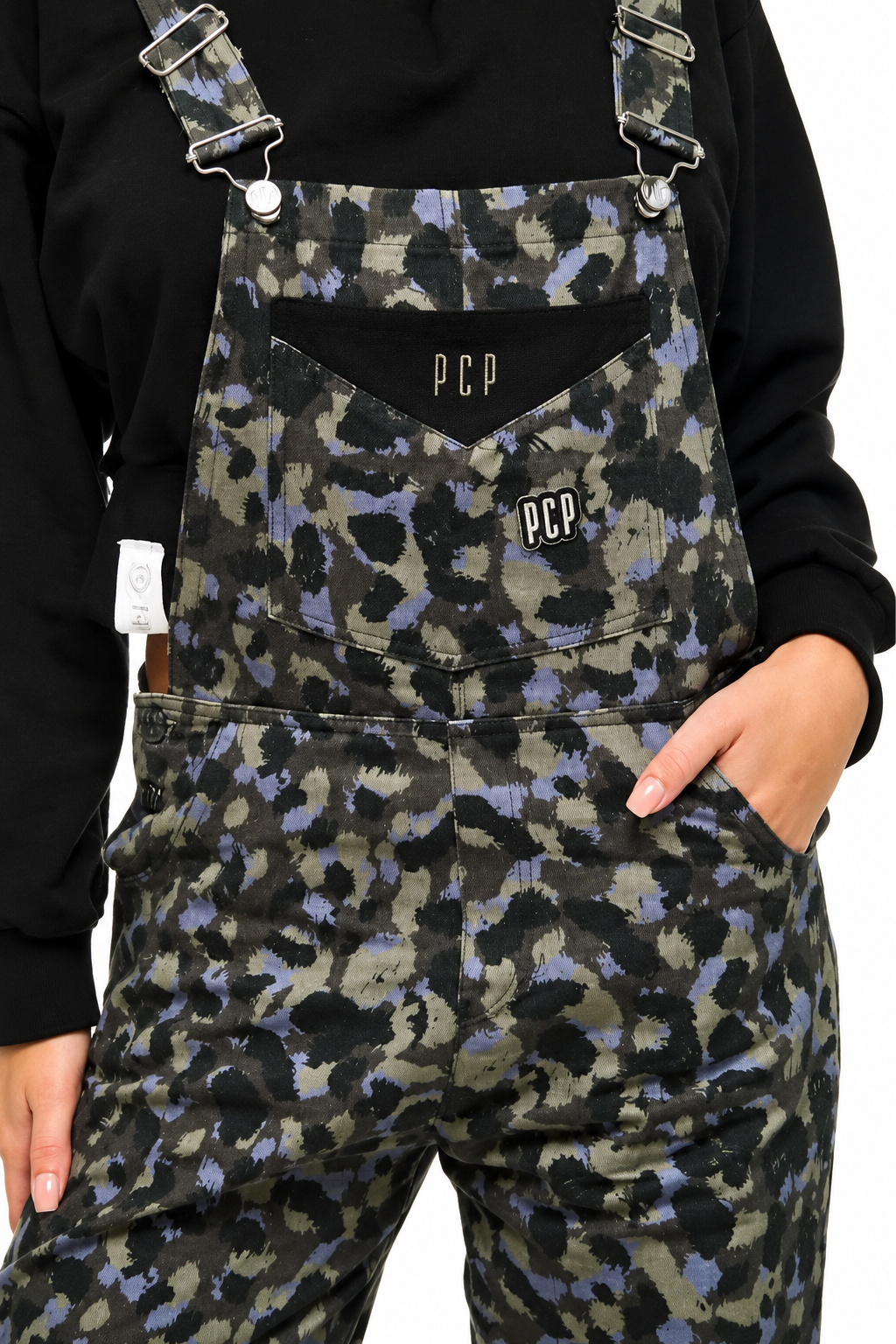 A close-up of a person wearing patterned overalls with a black long-sleeve top. The overalls feature a dark camouflage-style print in black, olive green, and muted purple, with a black chest pocket displaying the white 'PCP' logo.
