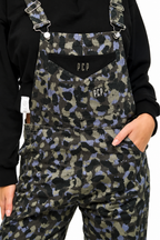 A close-up of a person wearing patterned overalls with a black long-sleeve top. The overalls feature a dark camouflage-style print in black, olive green, and muted purple, with a black chest pocket displaying the white 'PCP' logo.