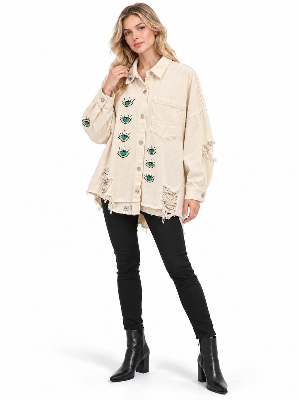 A woman modeling a distressed off-white denim jacket with green and black eye patches, paired with black skinny jeans and black ankle boots, on a white studio background.
