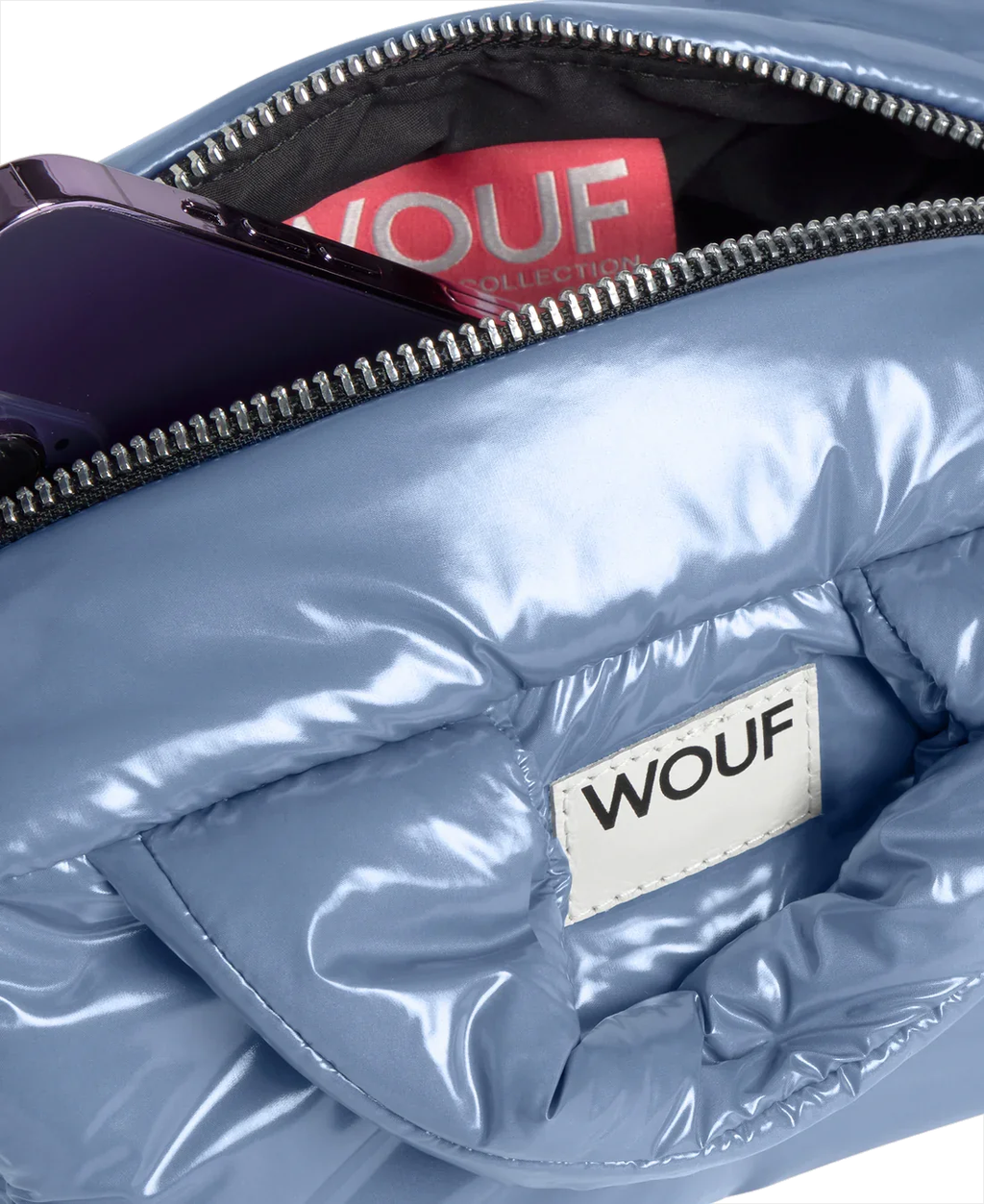 Close-up of a shiny light blue puffer bag with a silver zipper, showing a white sewn label that reads 'WOUF' and a red interior tag with the same brand.