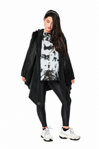 A woman modeling a black faux-fur hooded cape jacket over a black and white tie-dye t-shirt, black leggings, and white sneakers against a solid white background.