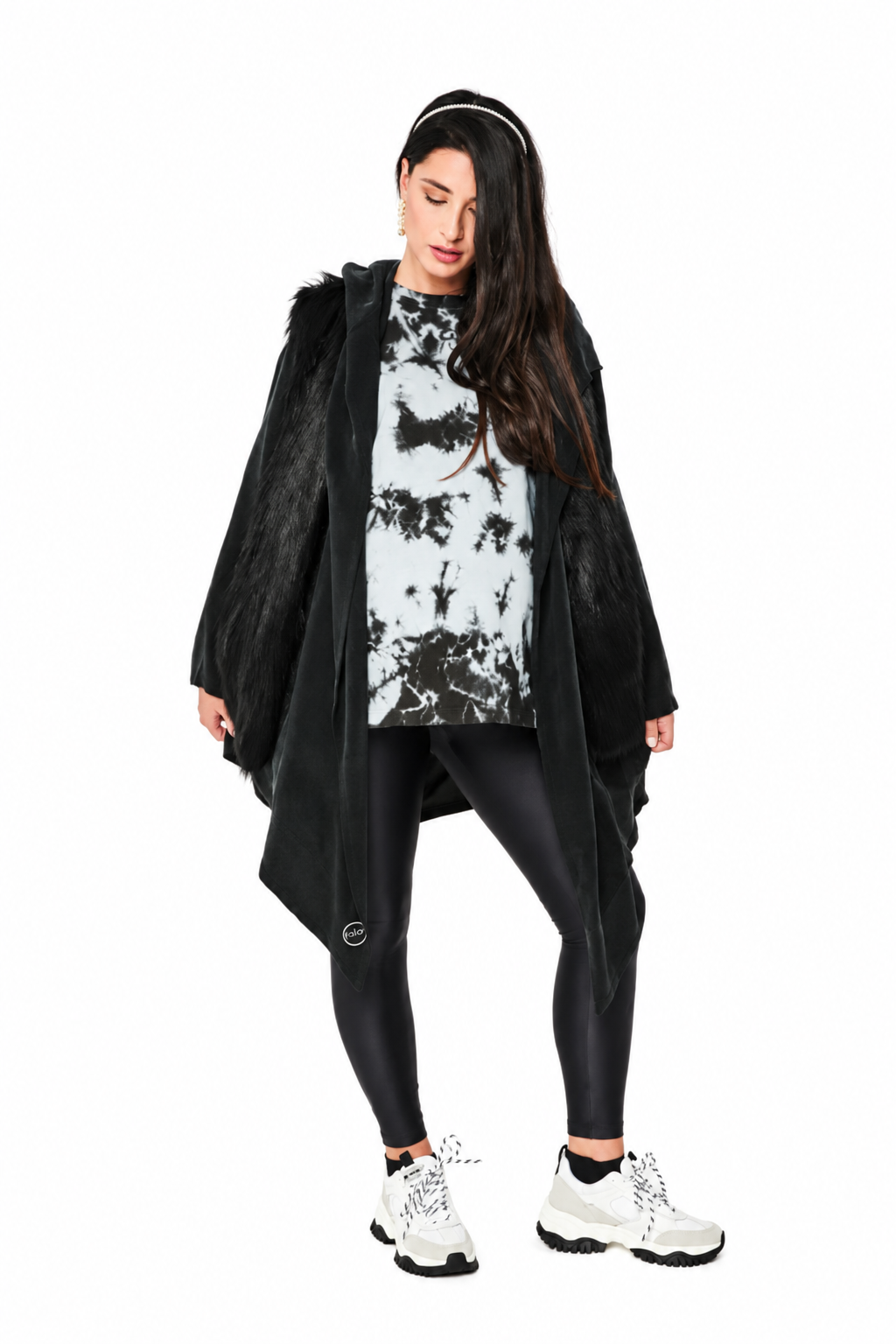 A woman modeling a black faux-fur hooded cape jacket over a black and white tie-dye t-shirt, black leggings, and white sneakers against a solid white background.