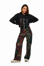 A model posing in a black cropped hoodie with colorful graphic details and high-waisted, wide-leg pants featuring a vibrant multicolor bohemian pattern, set against a plain white background.