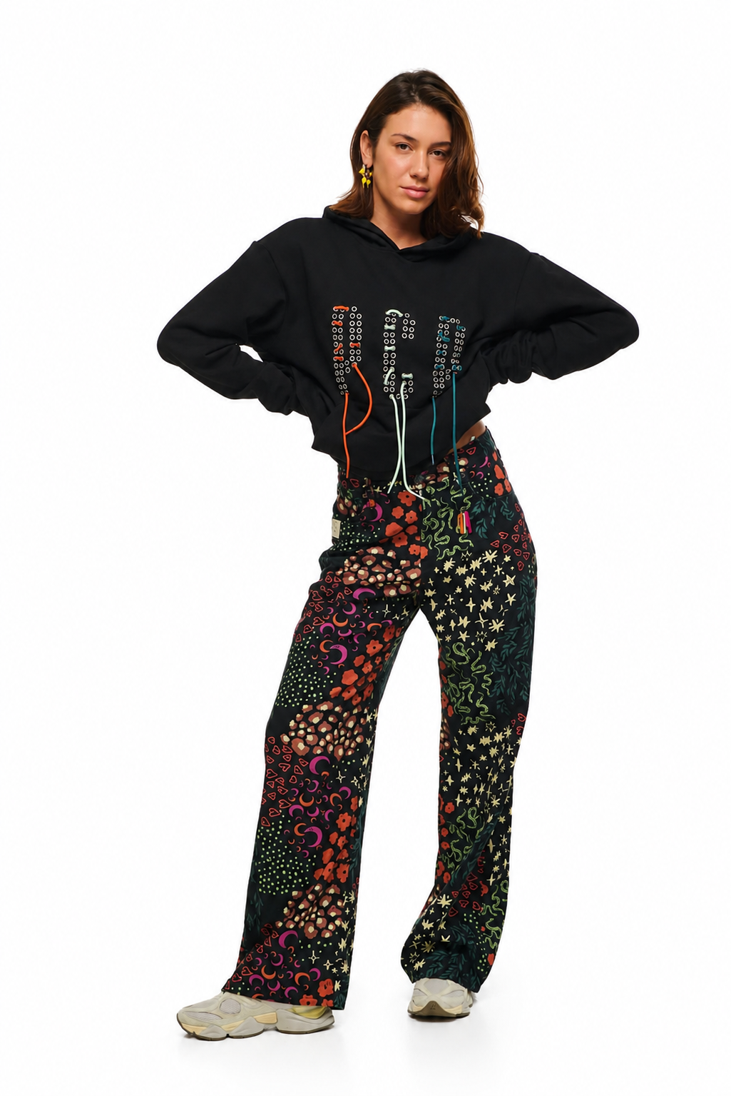 A model posing in a black cropped hoodie with colorful graphic details and high-waisted, wide-leg pants featuring a vibrant multicolor bohemian pattern, set against a plain white background.