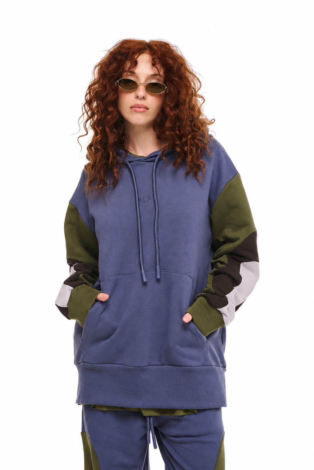 A model with curly red hair and sunglasses wearing a color-blocked hoodie and matching sweatpants. The hoodie features a slate blue body with olive green and white color-blocked sleeves and hood, set against a plain white background.