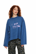 A model with curly red hair wearing a blue long-sleeve graphic sweatshirt and camouflage pants, standing against a plain white background.