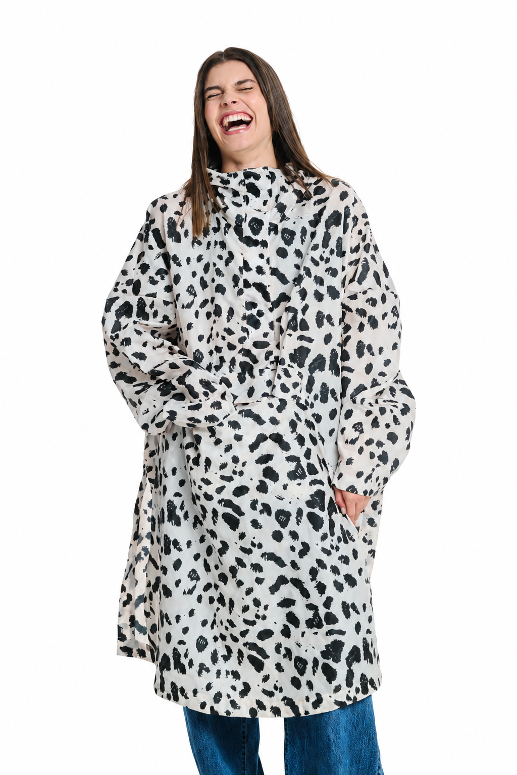 A woman laughing while wearing a high-neck, long-sleeved coat with an oversized black and white dalmatian-style animal print, paired with blue jeans, against a solid white background.