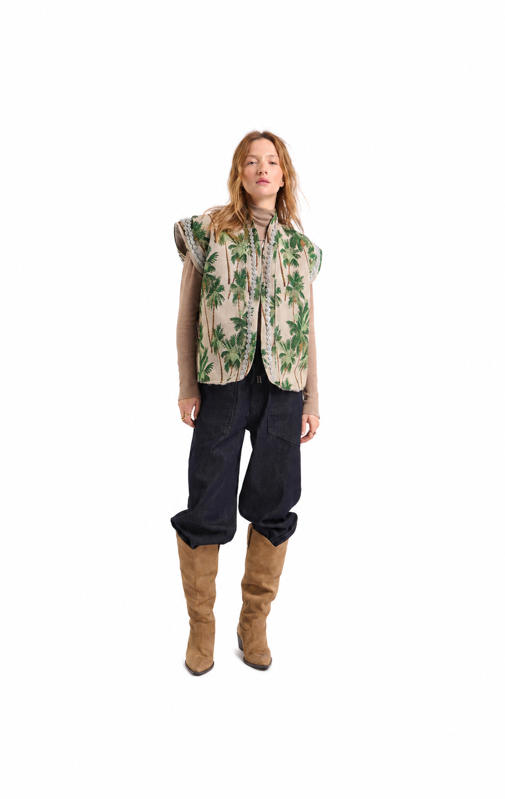 A model stands against a solid white background wearing a sleeveless beige vest with a green palm tree print, a beige long-sleeve shirt, dark wide-leg jeans, and tan knee-high boots.