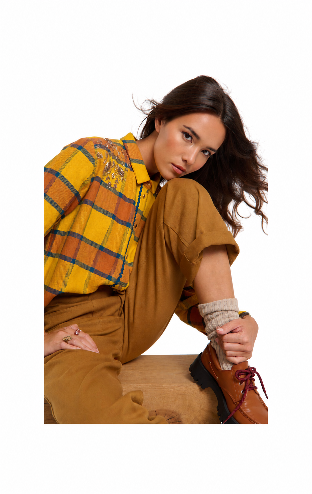 A woman posing in a yellow and orange plaid button-up shirt, brown trousers, beige knit socks, and brown leather shoes against a solid white background.