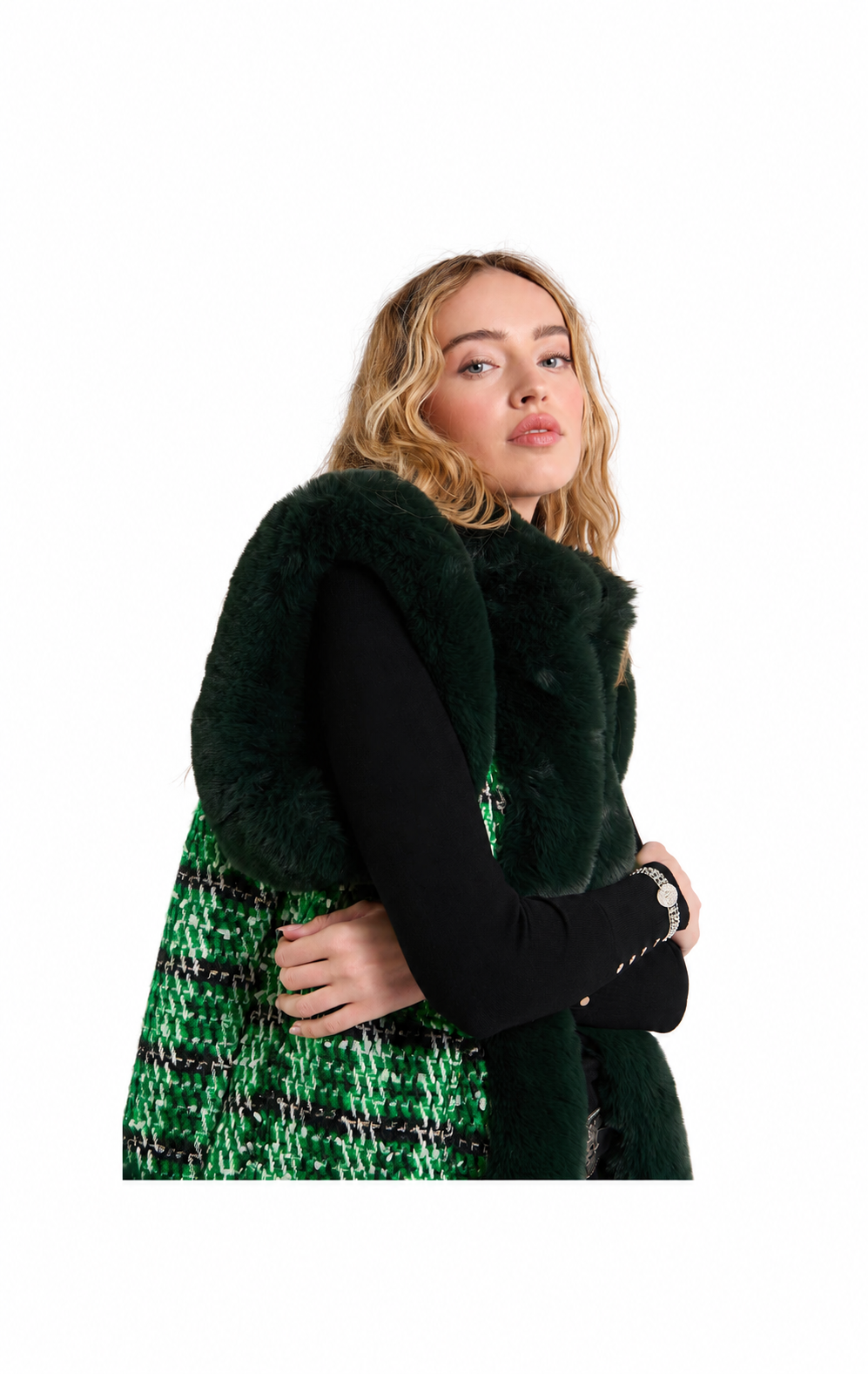 A woman modeling a dark green faux fur vest over a black long-sleeve top and a green and black tweed skirt, set against a plain white background.