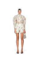 A model stands against a white studio background wearing a sheer, light beige floral long-sleeve shirt and matching shorts, with a patterned headscarf, a small woven handbag, and black flat sandals.