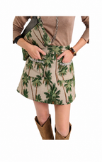 A model wearing a beige mini skirt with a green palm tree print, styled with a brown long-sleeve top and brown boots against a white background.
