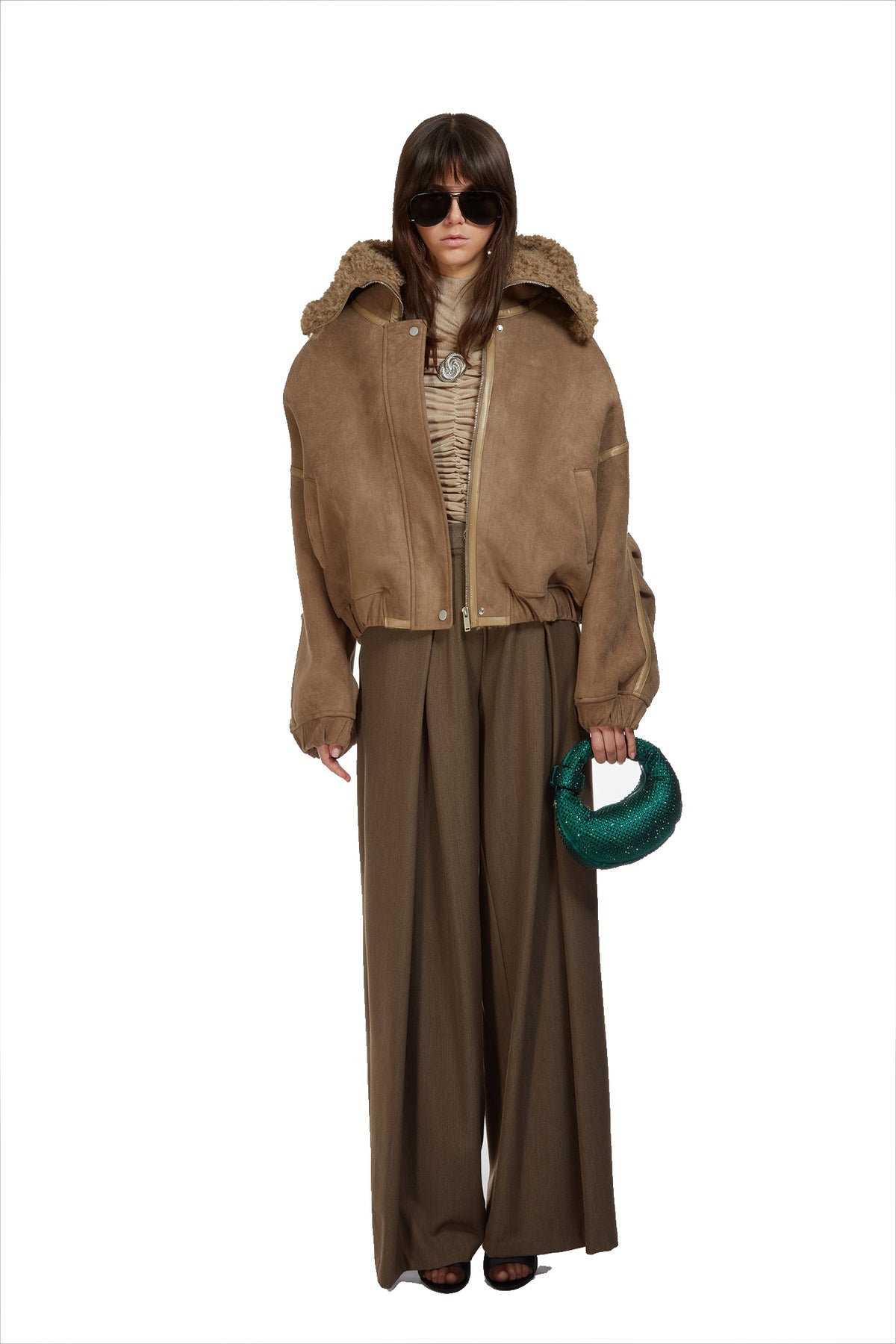 A model wearing a brown suede cropped jacket with a fur-trimmed collar, wide-leg brown pants, a patterned beige top, and sunglasses, holding a green textured ring against a plain white background.