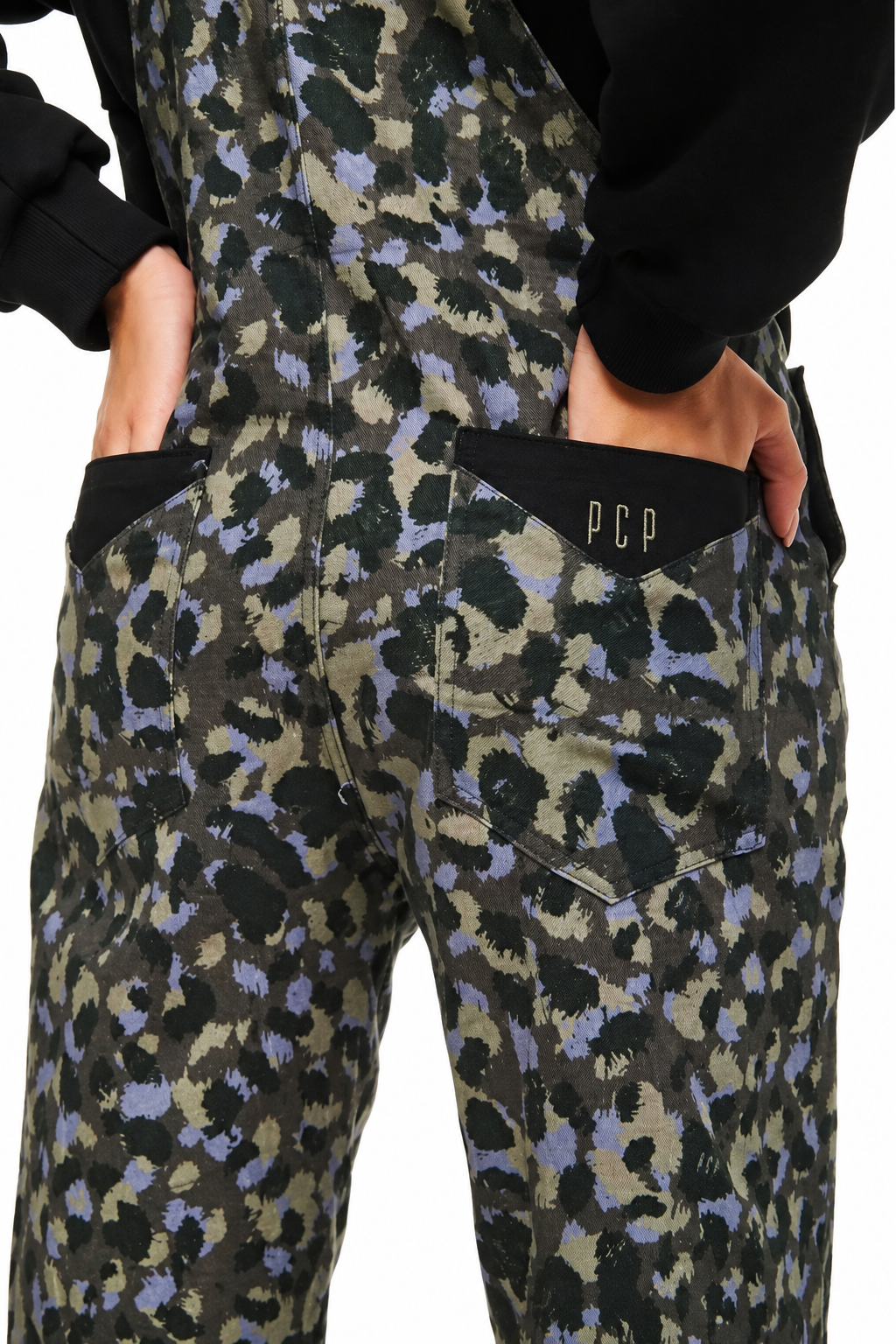Close-up back view of a person wearing overalls with an olive green, black, and lavender-purple leopard print pattern. The person is wearing a black sweatshirt, and the back pockets of the overalls are visible, with 'PCP' printed in gold on the right pocket.