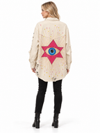 A woman shown from the back wearing a cream-colored distressed denim jacket with a large pink star and blue eye graphic on the back, paired with black pants and black boots, against a white background.