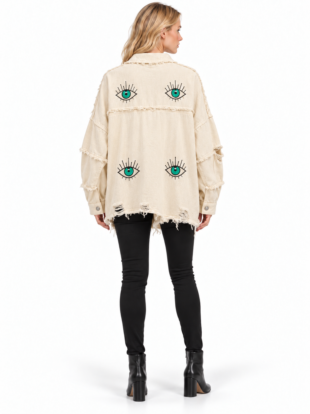 A woman shown from the back wearing a cream-colored, distressed denim jacket with embroidered green and blue eyes, paired with black skinny jeans and black ankle boots, against a plain white background.
