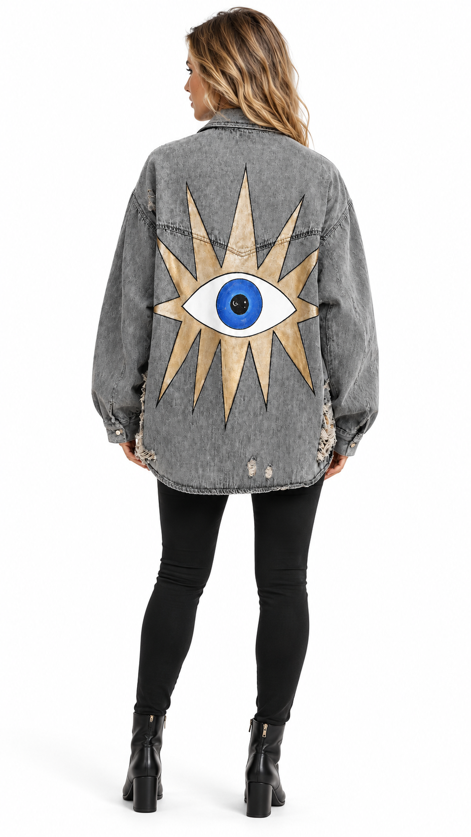 A woman viewed from the back wearing a gray washed denim jacket with a large gold and blue eye design on the back, paired with black leggings and black ankle boots, against a plain white background.