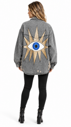 A woman viewed from the back wearing a gray washed denim jacket with a large gold and blue eye design on the back, paired with black leggings and black ankle boots, against a plain white background.