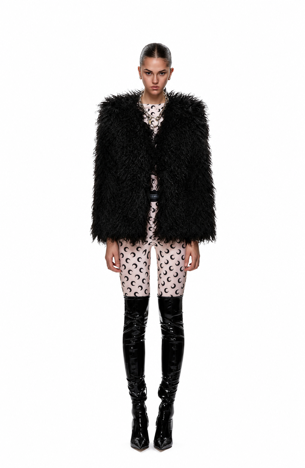 A model stands against a white studio background wearing a voluminous black shaggy faux-fur vest over a beige and black patterned top and shorts, paired with shiny black knee-high boots.