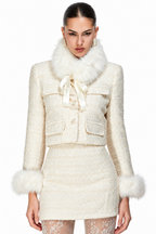 A model wearing a cream-colored tweed jacket and matching mini skirt set, both featuring fluffy faux-fur trim at the collar, cuffs, and hem, with a matching cream satin bow on the jacket.