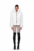 A model stands against a white studio background wearing a voluminous white faux-fur jacket with a large collar, paired with patterned pink and black cropped pants and glossy black over-the-knee boots.