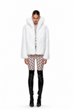 A model stands against a white studio background wearing a voluminous white faux-fur jacket with a large collar, paired with patterned pink and black cropped pants and glossy black over-the-knee boots.