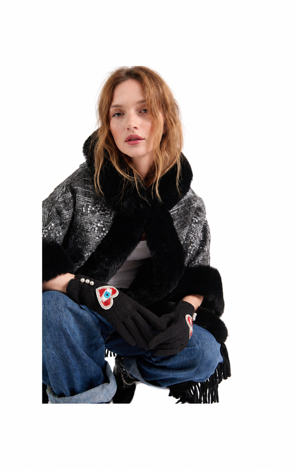 A woman wearing a black and gray patterned shawl with black fur trim, black knit gloves, and blue jeans, posed against a solid white background.