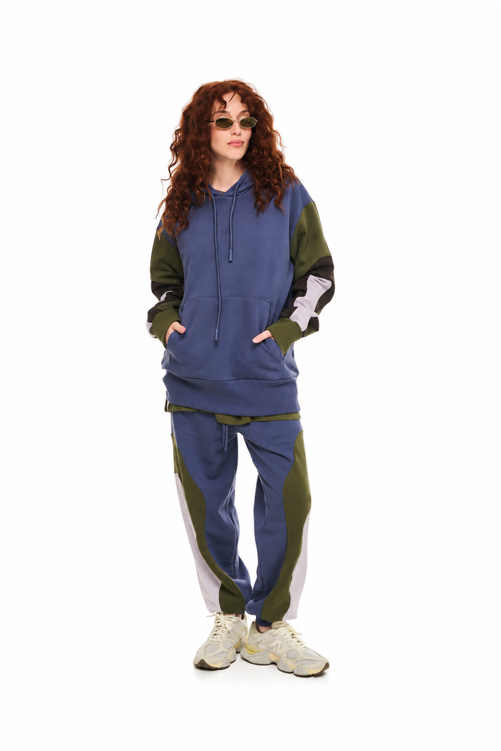 A model with curly red hair wearing a color-blocked hoodie and matching sweatpants set in blue, olive green, and white, paired with beige sneakers, on a solid white background.