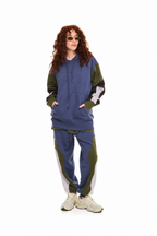A model with curly red hair wearing a color-blocked hoodie and matching sweatpants set in blue, olive green, and white, paired with beige sneakers, on a solid white background.