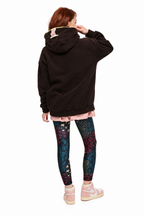 A model shown from the back wearing a black oversized hoodie with a pink bunny ear detail on the hood, a light pink pleated skirt, patterned leggings, and pink sneakers, set against a plain white background.